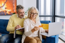 Ein Rentner-Ehepaar, das fröhlich auf der Couch mit dem Laptop und einer Kreditkarte Online-Banking betreibt.