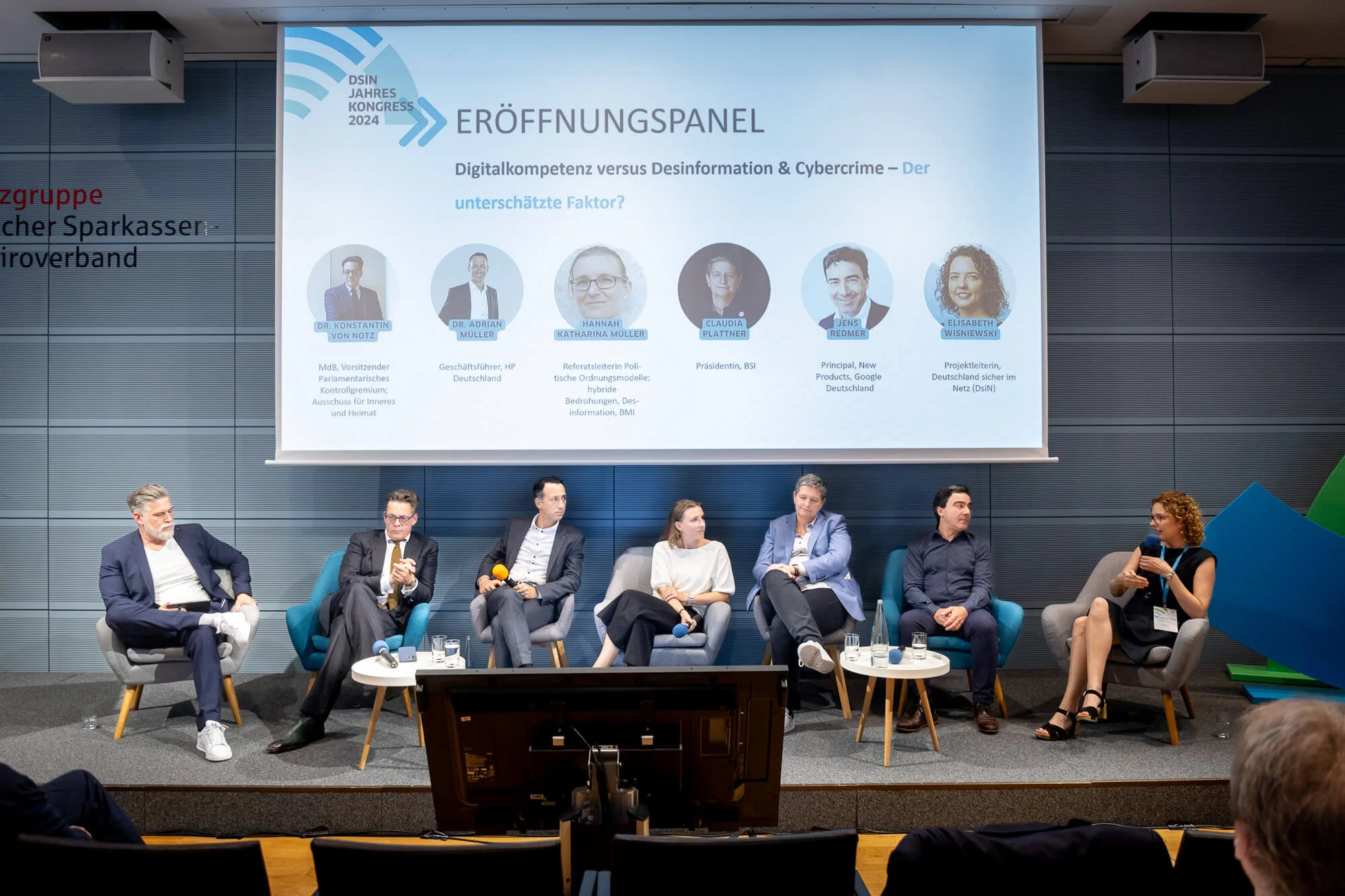 Eine Diskussionsrunde mit sieben Personen sitzt auf einer Bühne beim DsiN Jahreskongress 2024. Auf der Leinwand darüber steht Eröffnungspanel Digitalkompetenz versus Desinformation und Cybercrime – Der unterschätzte Faktor?