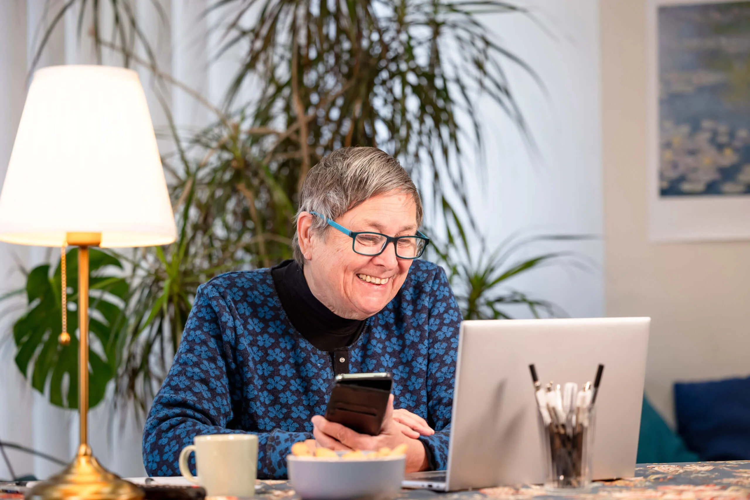 Eine Frau mit grauen Haaren sitzt lächelnd vor einem Laptop. Sie hat ein Smartphone in der Hand.