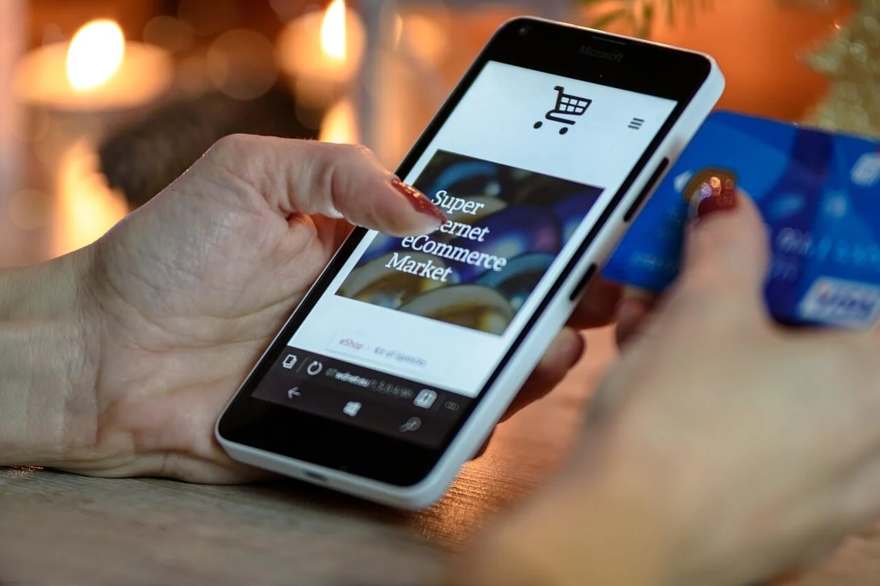 Es sind Hände zu sehen und in der einen Hand liegt ein Smartphone auf dessen Display ein Onlineeinkaufsverfahren zu sehen ist und in der anderen Hand eine Kreditkarte